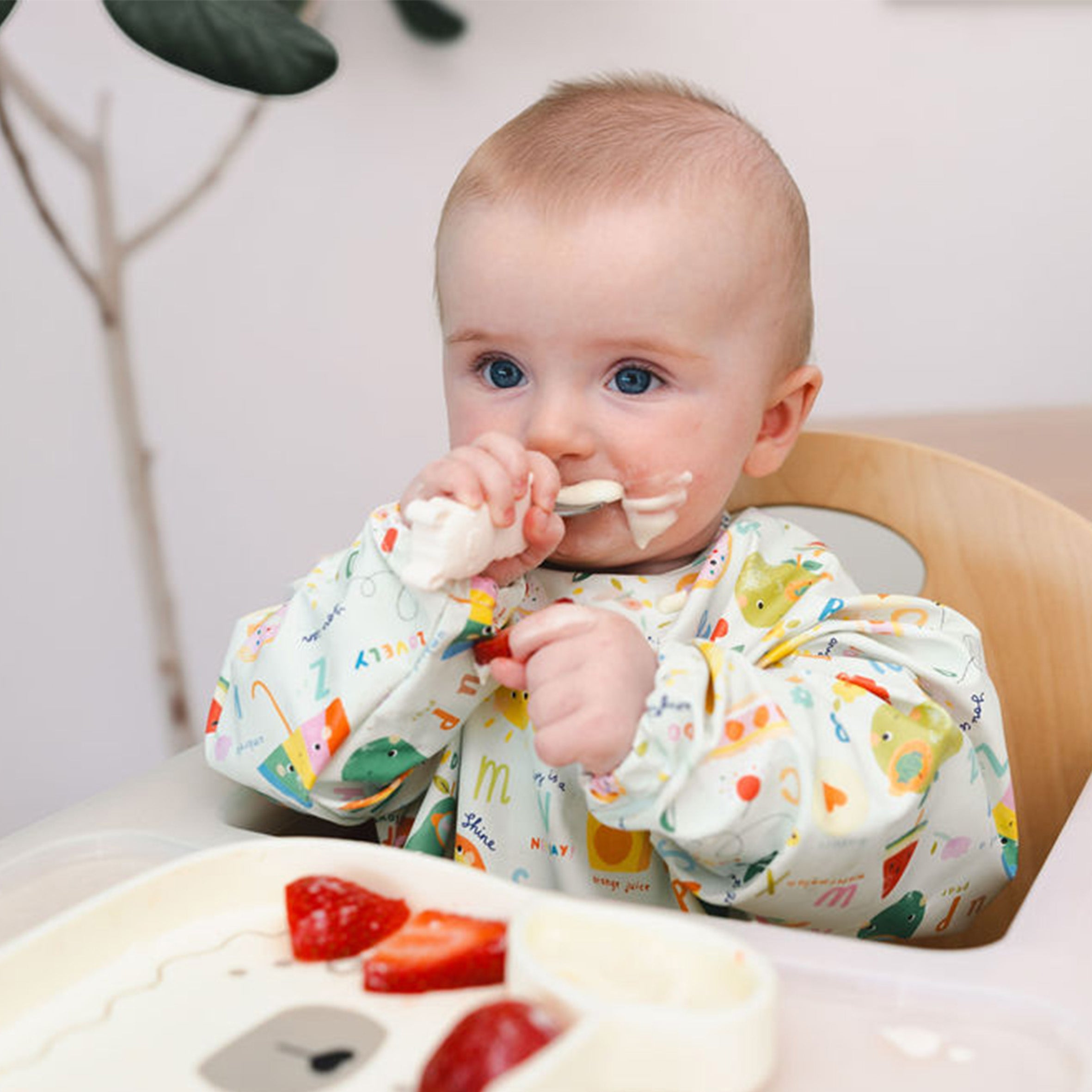 Baby in a high chair eating with a Lane Llama Learning Utensil spoon, wearing a Long Sleeve bib