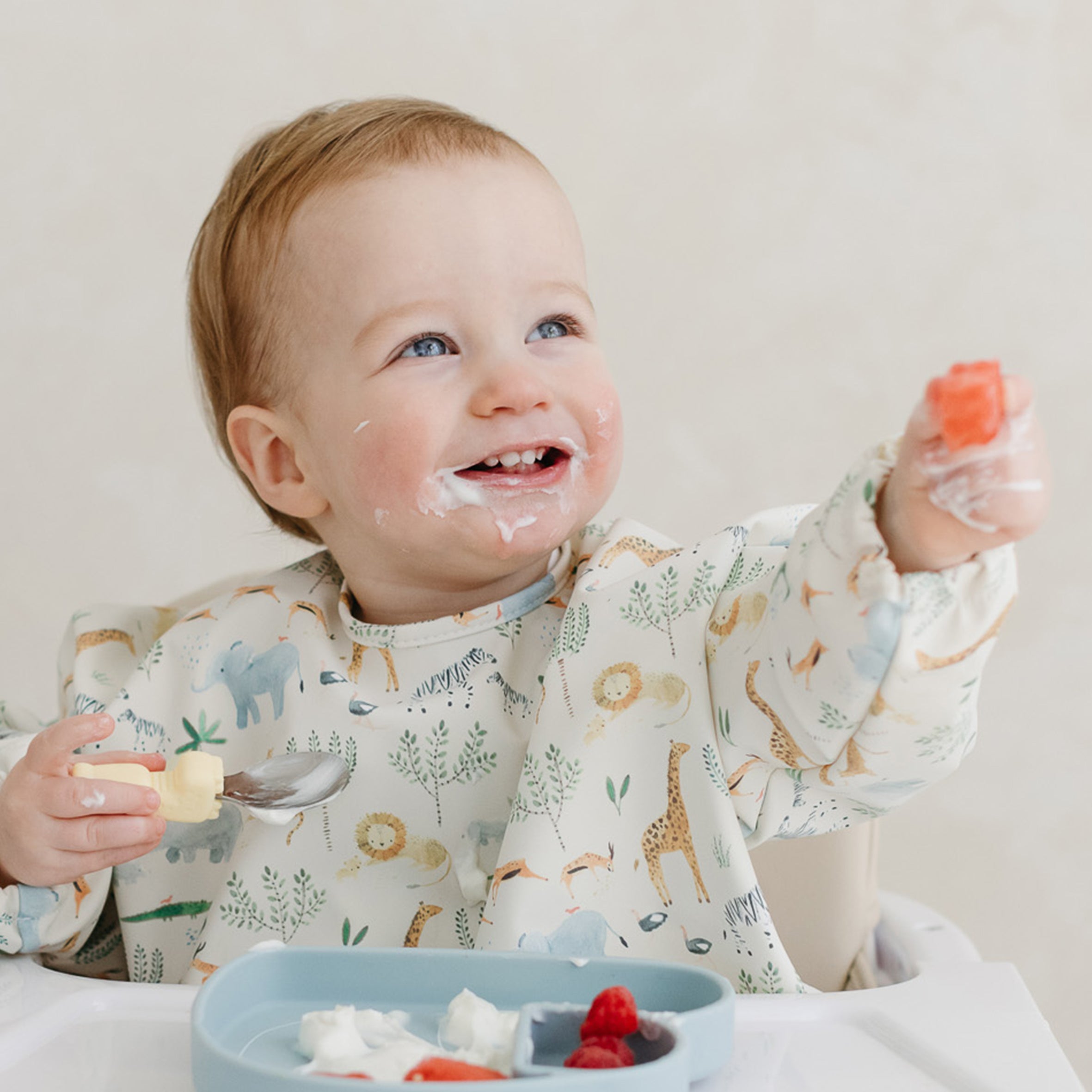 Baby in highchair wearing a Safari Jungle Long Sleeve Bib eating yoghurt with a Lane Llama Learning Utensils
