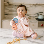 Baby sitting on a kitchen counter wearing a Sea Shells Long Sleeve Bib and scattered snacks