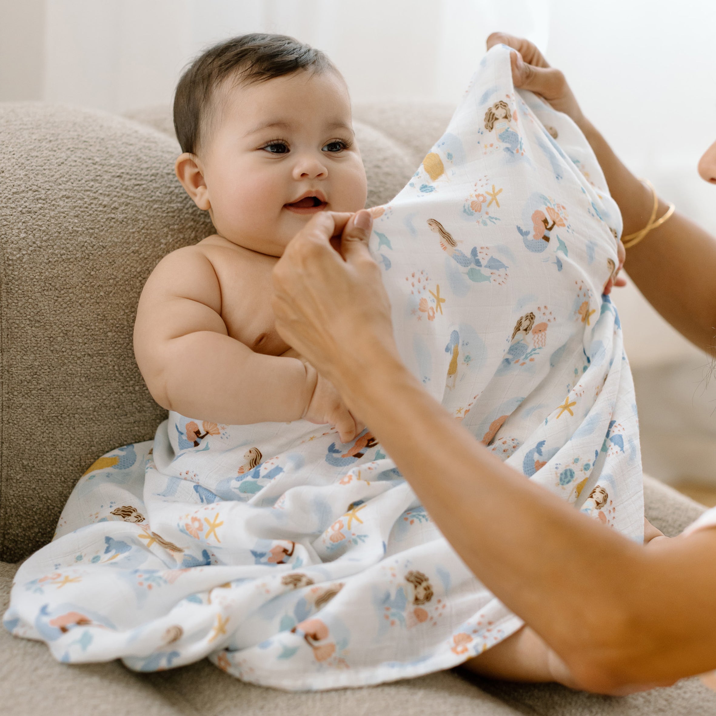 Baby being swaddled with a Mermaids Muslin Swaddle by an adult on a couch.