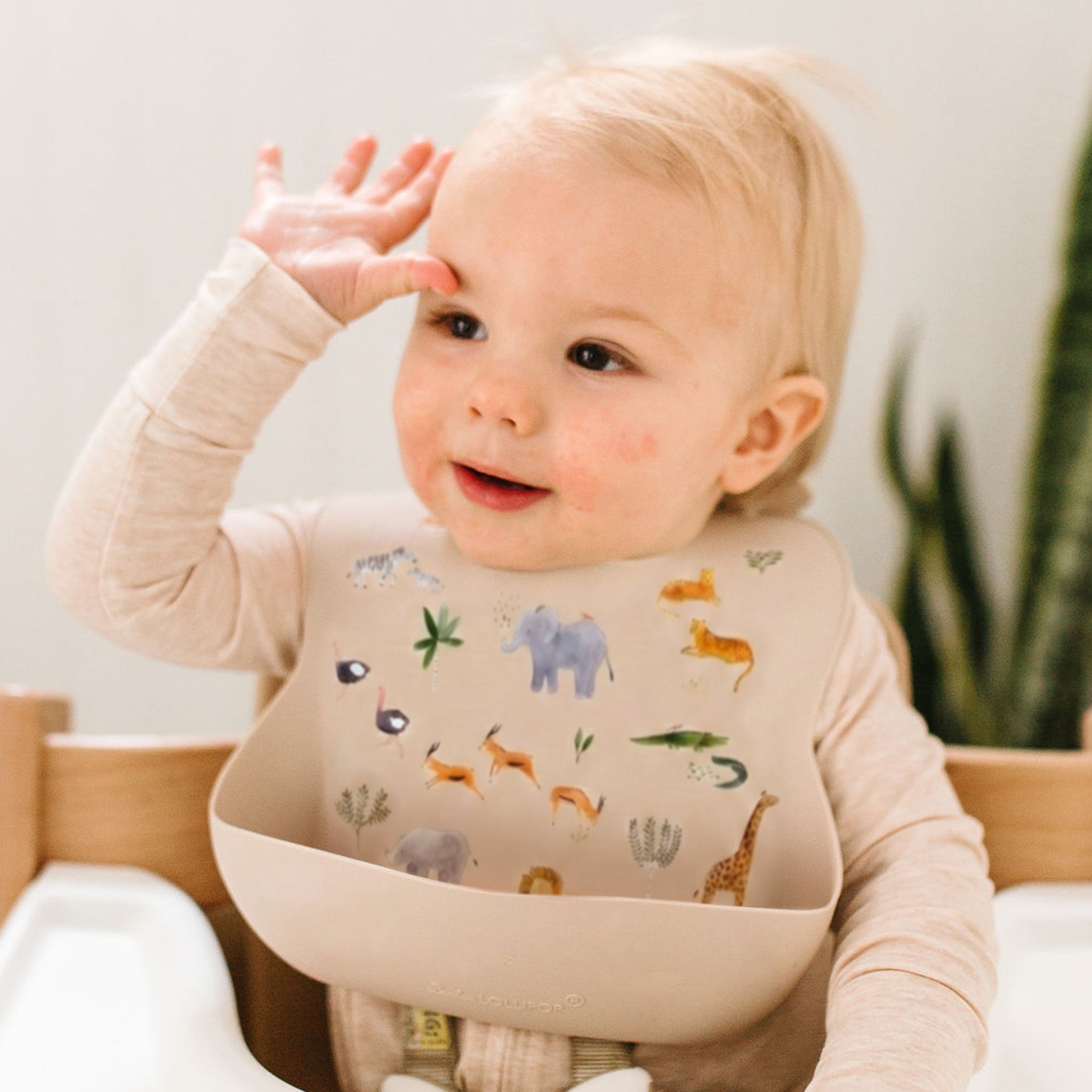 A baby wearing a beige long-sleeve shirt and a Safari Jungle silicone bib with colorful animal illustrations, sitting in a wooden high chair.
