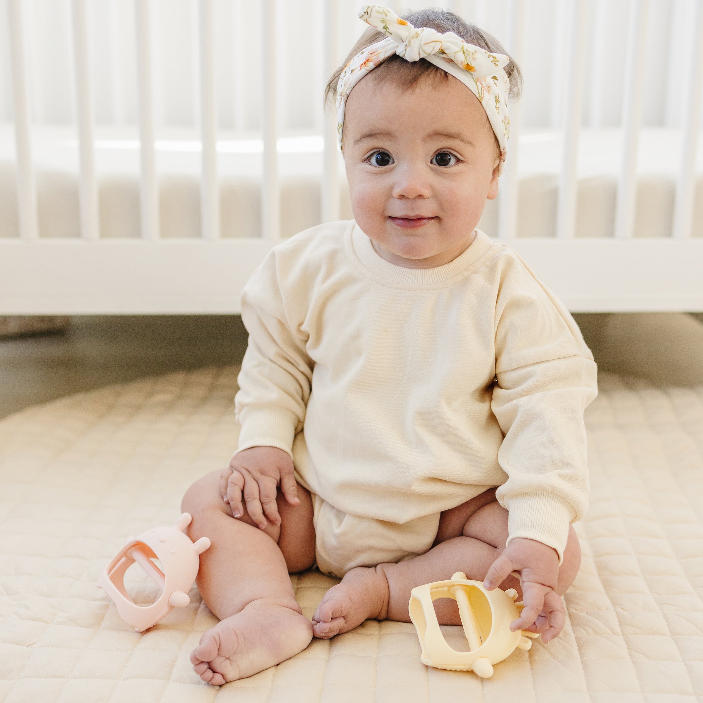 Baby swearing a white outfit and headband, holding toys.