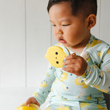 A young child wearing a light blue long-sleeve shirt with a yellow lemon print is holding and biting The Lemon Teething Clip teether toy.
