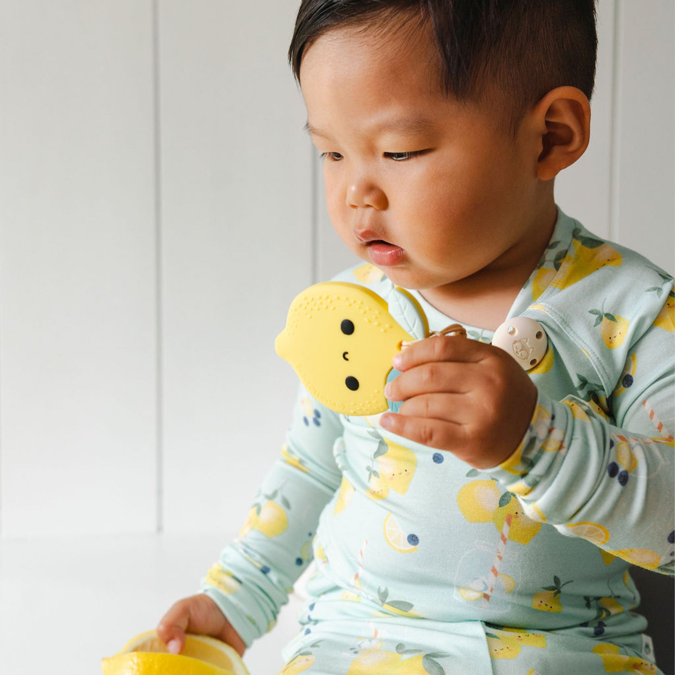 A young child wearing a light blue long-sleeve shirt with a yellow lemon print is holding and biting The Lemon Teething Clip teether toy.