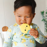 A young child wearing a light blue long-sleeve shirt with a yellow lemon print is holding and biting The Lemon Teething Clip teether toy.
