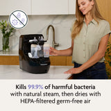 A woman uses the Baby Brezza Bottle Washer Pro® - All Black on her kitchen counter. It kills 99.9% of harmful bacteria with natural steam, dries bottles with HEPA-filtered air, and offers automatic cleaning for peace of mind. Bacteria icon crossed out.