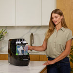 A woman places baby bottles and accessories into the all-black Baby Brezza Bottle Washer Pro® by Baby Brezza on a white kitchen countertop, with cabinets and a sink in the background.
