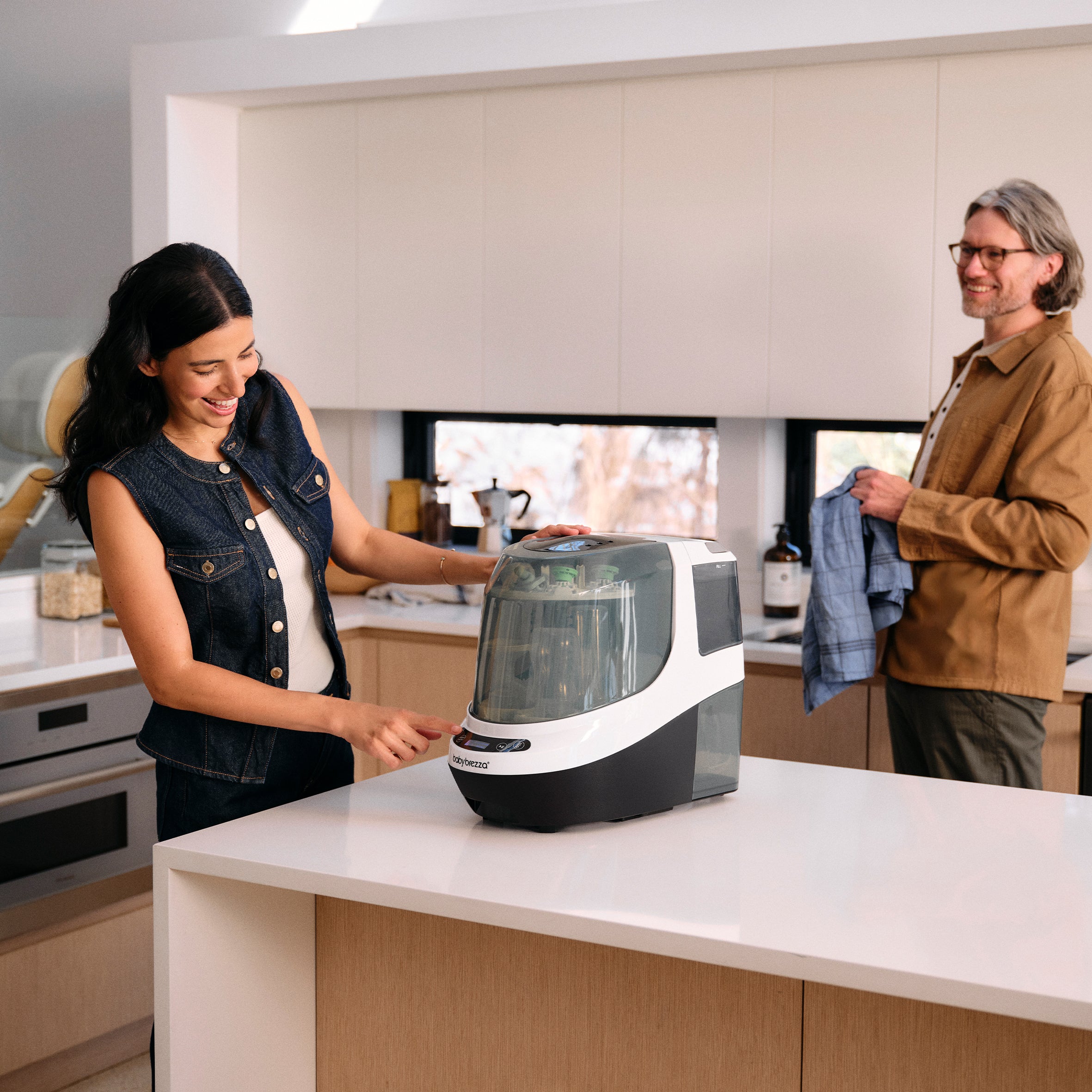 A woman presses the start button on a white and grey PRE-LOVED: Baby Brezza Bottle Washer Pro® - Classic White sitting on a kitchen counter, while a man stands nearby holding a towel.