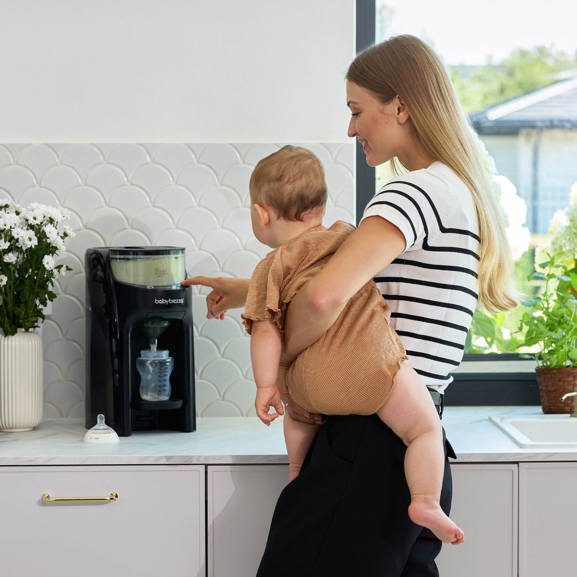A woman holds a baby who touches the control panel of a Baby Brezza Formula Pro Advanced on a kitchen counter.