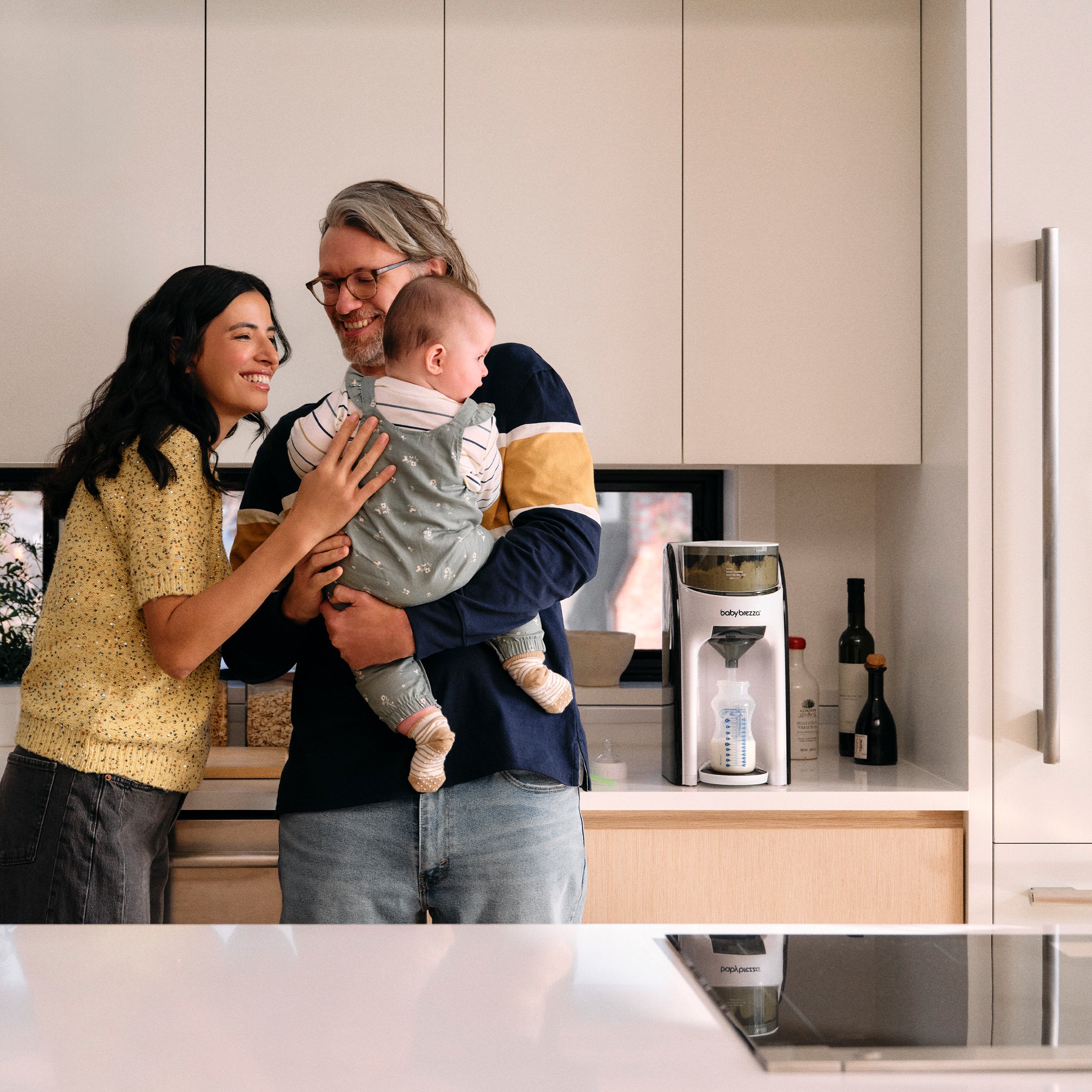 A smiling couple stands in a kitchen holding a baby next to a Baby Brezza Formula Pro Advanced Bundle on the countertop.