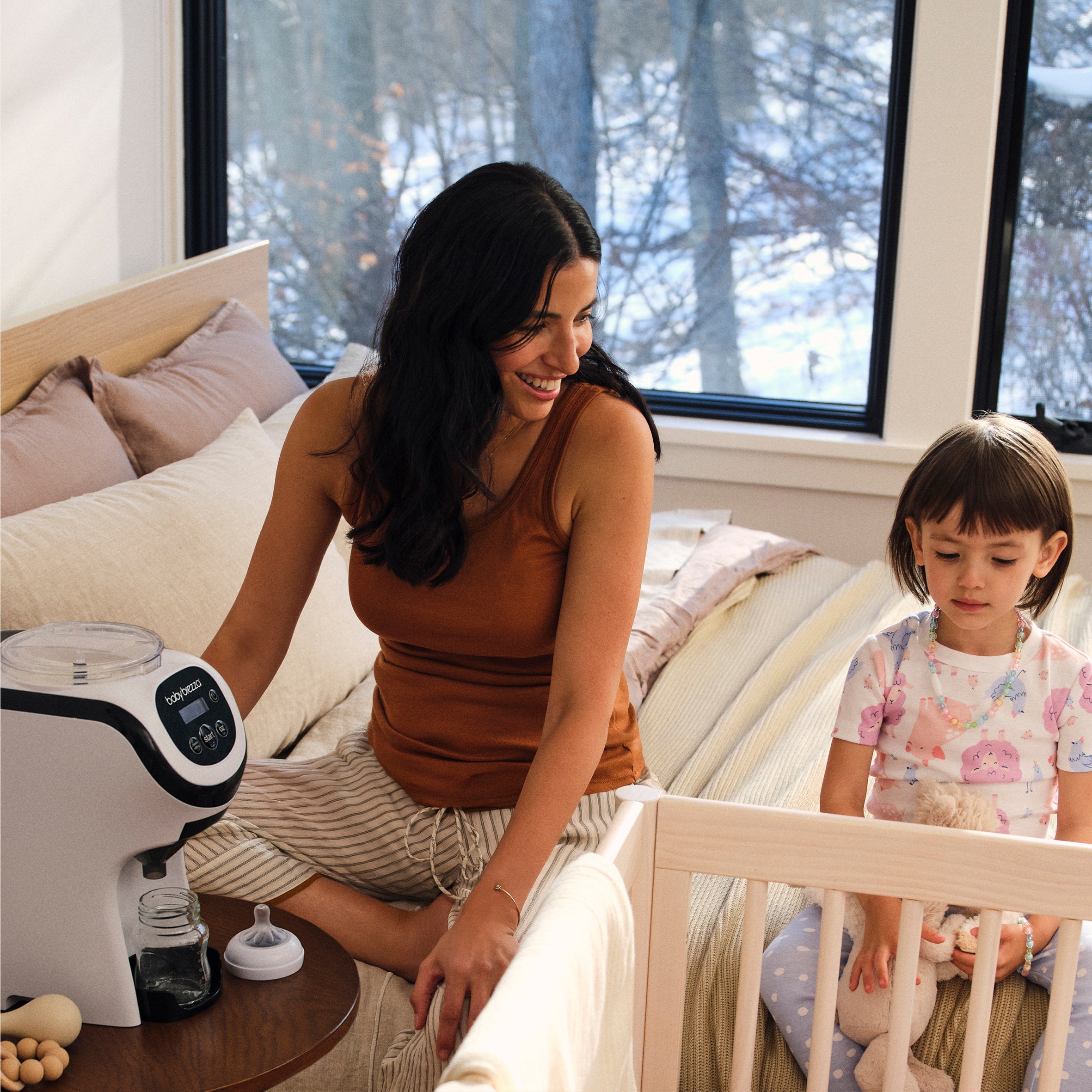 A woman sits on a bed next to a Baby Brezza Formula Pro Mini while a young child sits in a crib nearby.