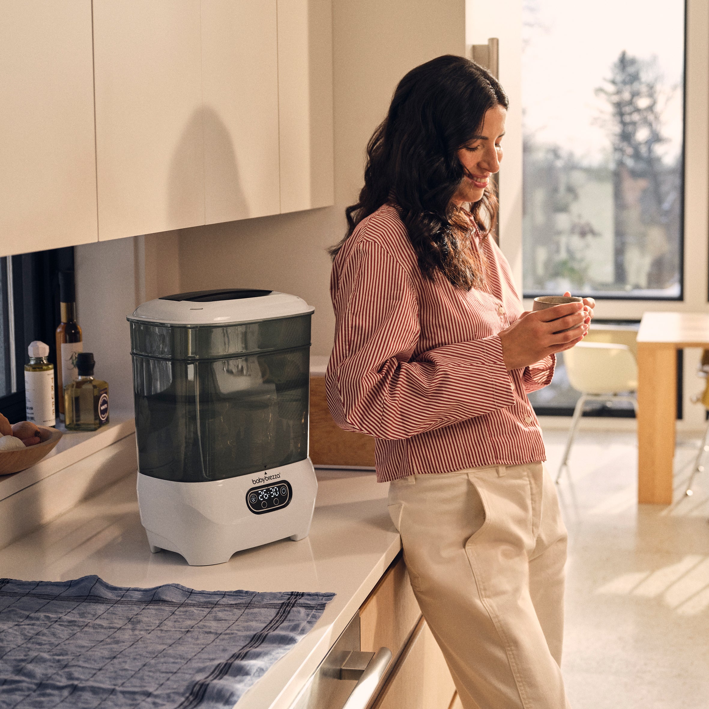 A woman leans against a kitchen counter next to a classic white Baby Brezza Steriliser Dryer Advanced.