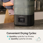 A person uses a Baby Brezza SuperQuick Steriliser Dryer on the kitchen counter. The display shows drying settings. Text: “Convenient Drying Cycles—QuickDry (5 & 10 mins), QuietDry (25 mins)” with checkmarks and a clock icon.
