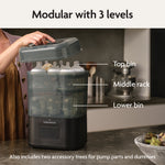 On a kitchen counter, someone lifts the lid of the Baby Brezza SuperQuick Steriliser Dryer, a modular 3-level unit with top bin, middle rack, and lower bin. It includes two accessory trees for pump parts and dummies and features 4-in-1 functionality.