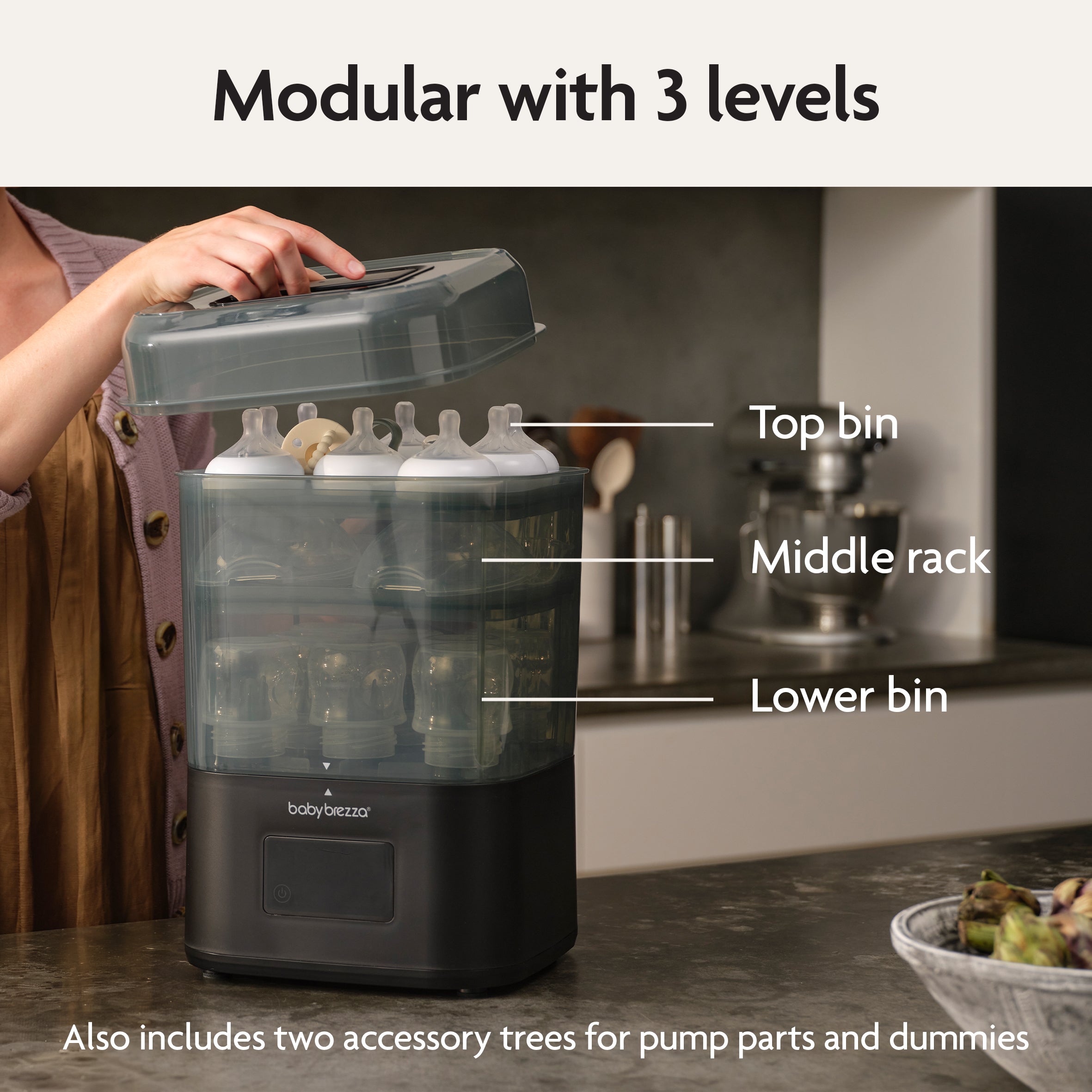 On a kitchen counter, someone lifts the lid of the Baby Brezza SuperQuick Steriliser Dryer, a modular 3-level unit with top bin, middle rack, and lower bin. It includes two accessory trees for pump parts and dummies and features 4-in-1 functionality.