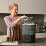 A woman smiles as she opens the lid of a Baby Brezza SuperQuick Steriliser Dryer, filled with several baby bottles, on her kitchen counter.