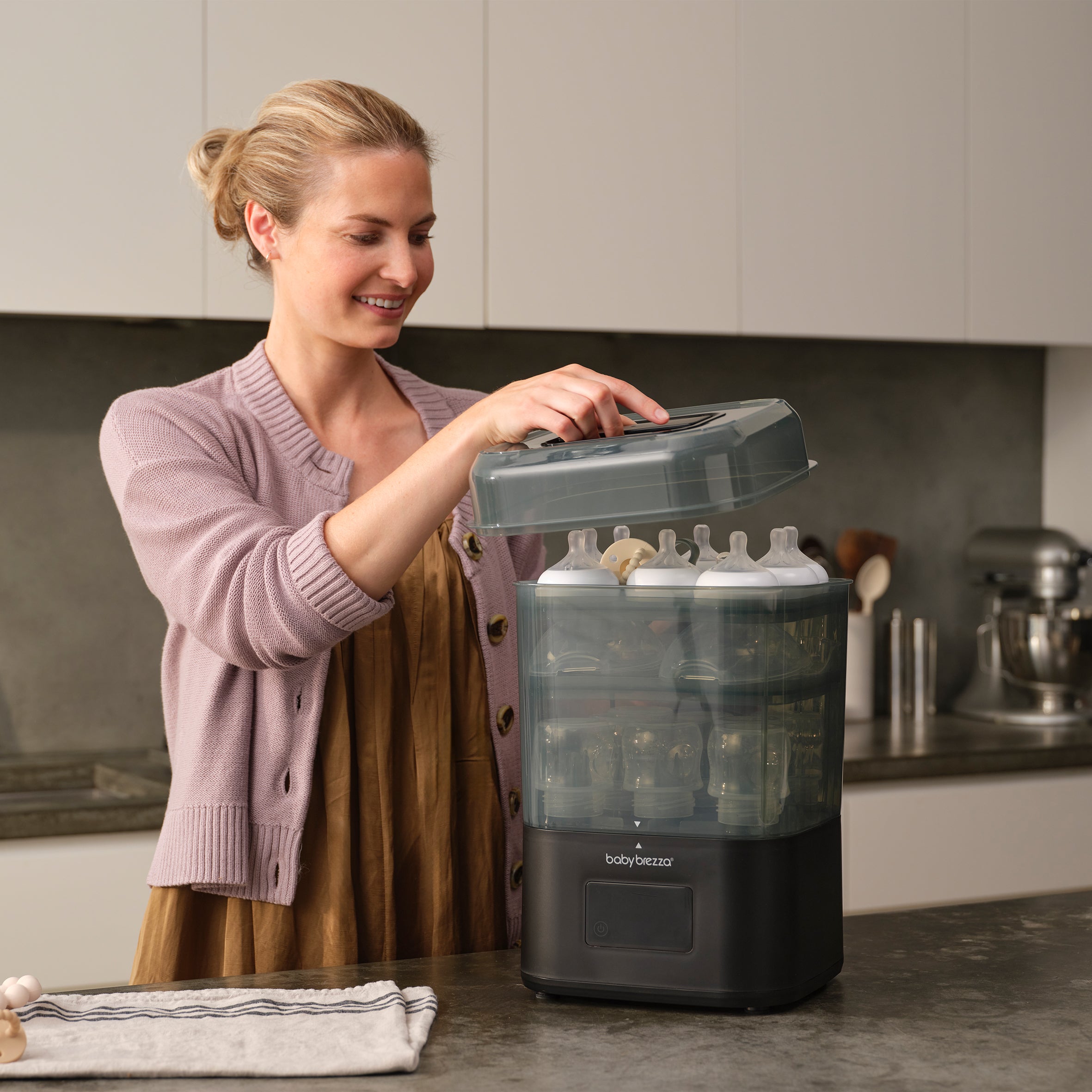 A woman smiles as she opens the lid of a Baby Brezza SuperQuick Steriliser Dryer, filled with several baby bottles, on her kitchen counter.