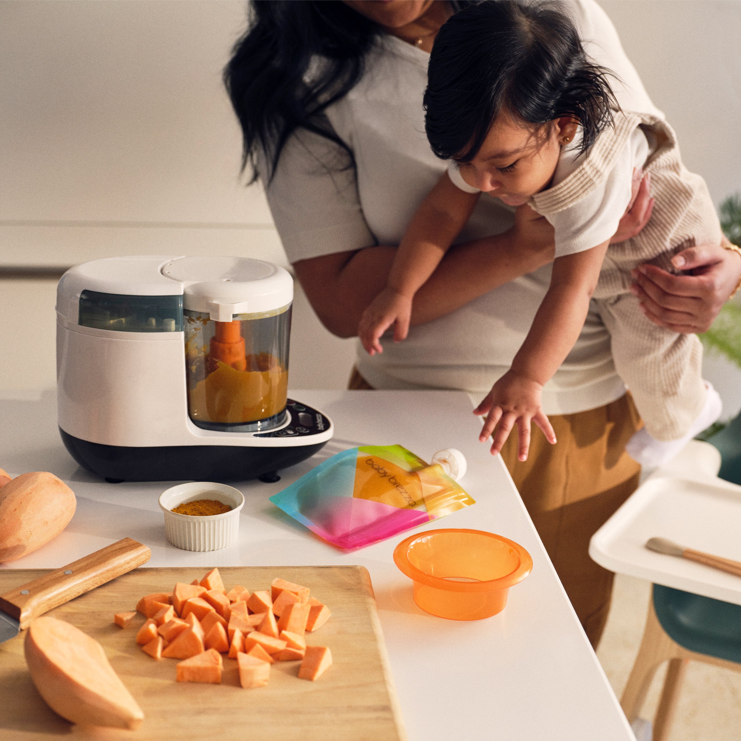 A person holding a baby stands at a counter next to a Baby Brezza Food Maker Deluxe, a wooden cutting board with chopped sweet potatoes, a small bowl of spice, and a colorful baby food pouch.