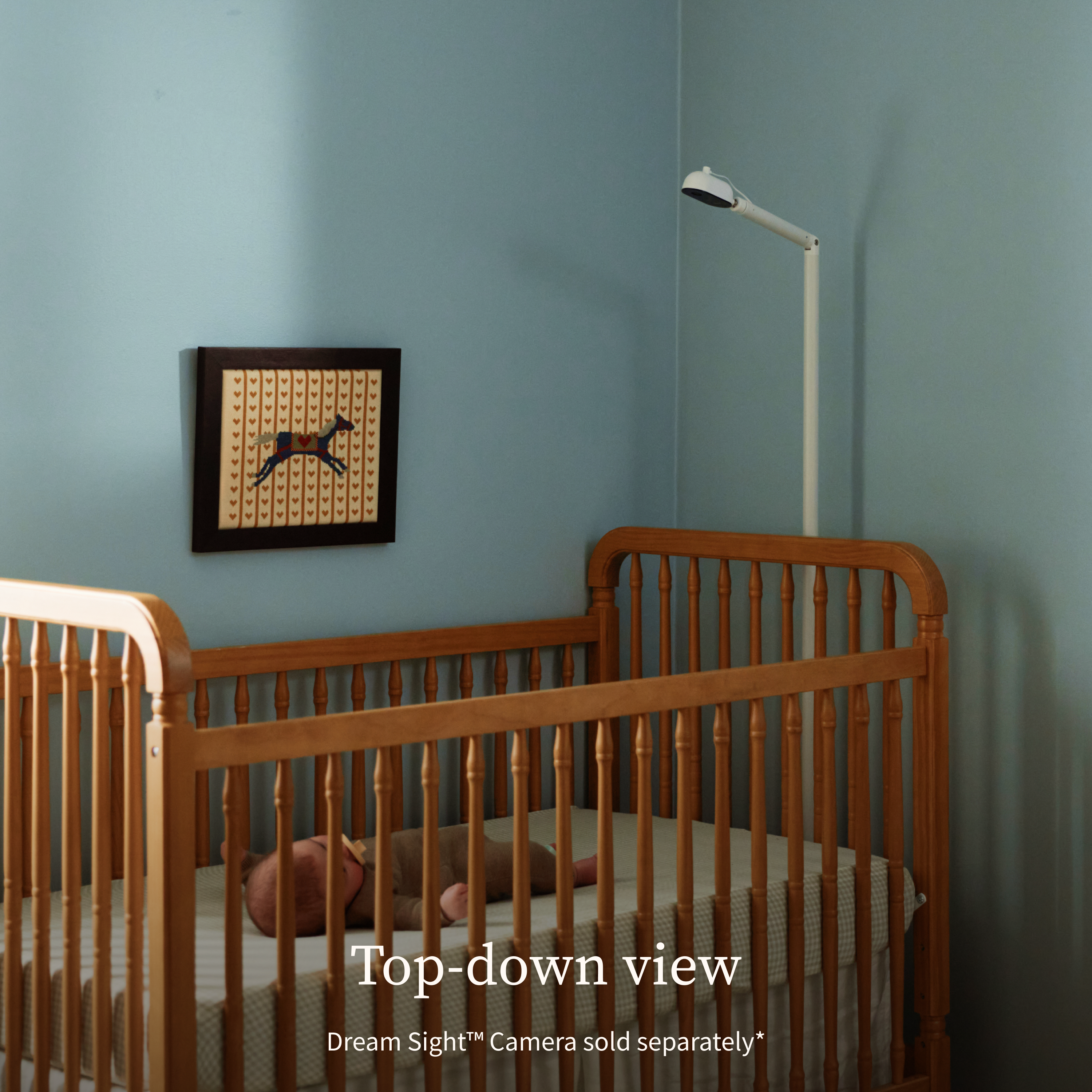 A tall, white Owlet Dream Sight™ Camera Stand is positioned behind a wooden crib, with the baby camera angled downward to monitor an infant lying inside.