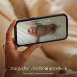 A hand holds a smartphone displaying a baby monitor app with vitals, while an Owlet Dream Sight™ Camera Stand holds the device over a baby resting on a crib mattress.