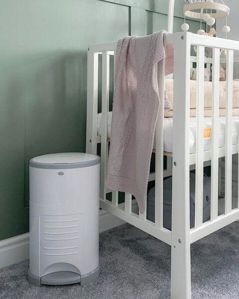A white crib with a light pink blanket draped over the side stands beside the Korbell Eco Nappy Bin in Pure White by Korbell, which offers hands-free nappy disposal and odour control, set on a gray carpet in a nursery with light green walls and a hanging mobile.