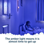 A child sits in bed under blue light, reading a book while an amber nightlight glows on the wall beneath Rockit’s Rising Star – The Rewarding Sleep Clock. Text reads: The amber light means it’s almost time to get up—thanks to this kids’ sleep trainer, mornings are easier for everyone.