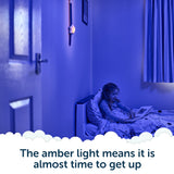 A child sits in bed under blue light, reading a book while an amber nightlight glows on the wall beneath Rockit’s Rising Star – The Rewarding Sleep Clock. Text reads: The amber light means it’s almost time to get up—thanks to this kids’ sleep trainer, mornings are easier for everyone.