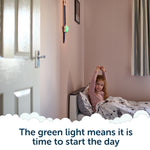 A young girl sits up and stretches in bed as a green light glows on the wall, signaling her children wake up clock; text below reads, The green light means it is time to start the day.