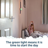 A young girl sits up and stretches in bed as a green light glows on the wall, signaling her children wake up clock; text below reads, The green light means it is time to start the day.