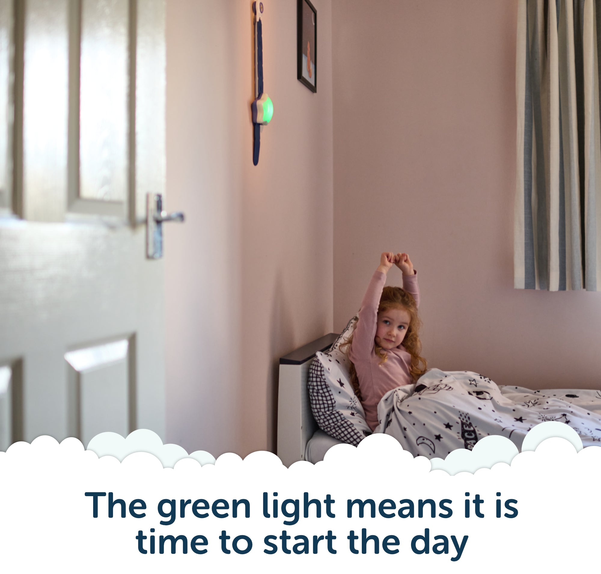 A young girl sits up and stretches in bed as a green light glows on the wall, signaling her children wake up clock; text below reads, The green light means it is time to start the day.